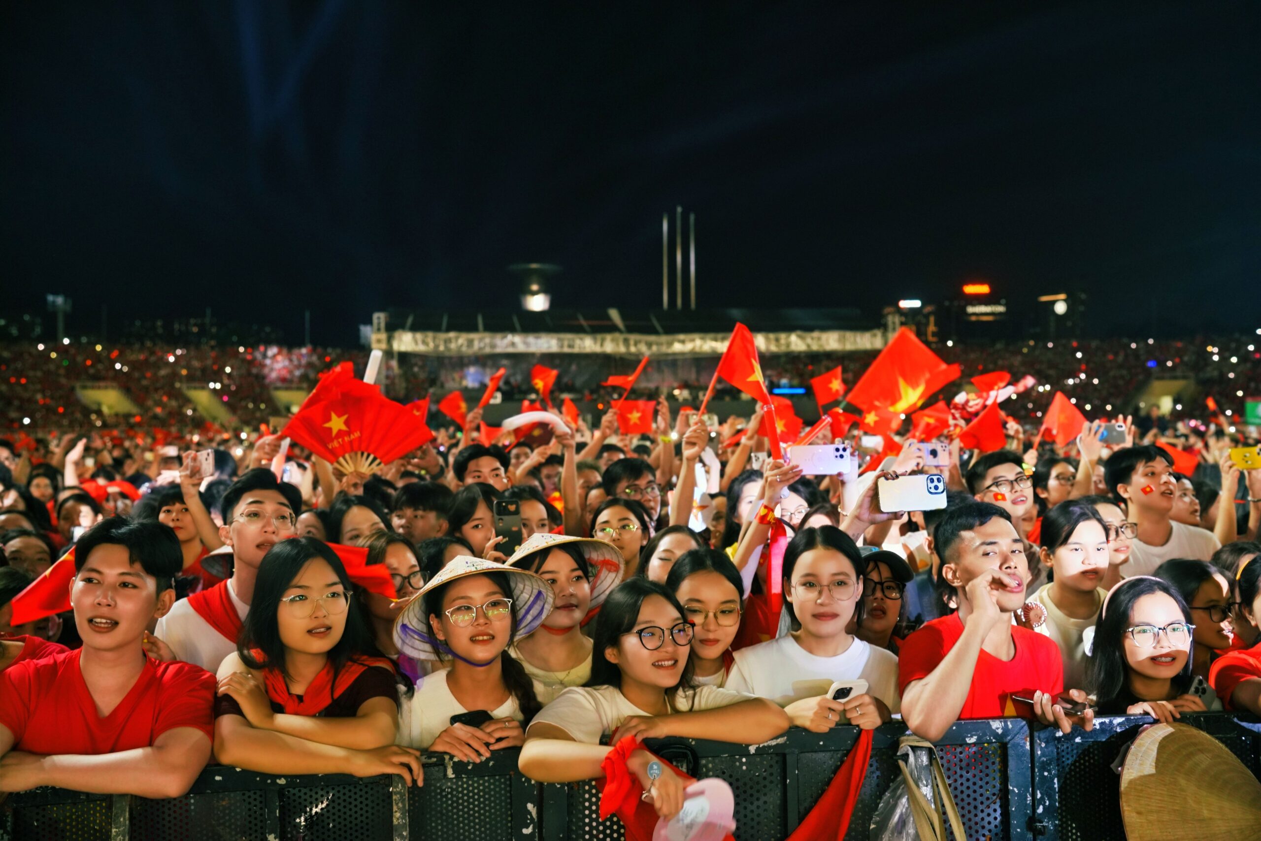 Excited crowd waving flags at a nighttime live event, representing fans gathered to celebrate their favorite stars.